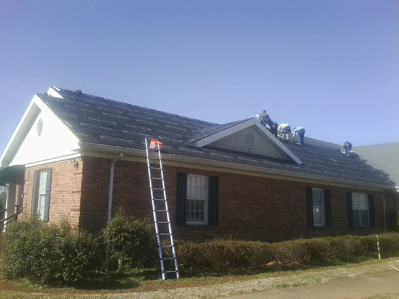 Crew tearing shingles off a brick ranch roof with a ladder set — Emergency Roof Repair work by our crew in Delavan